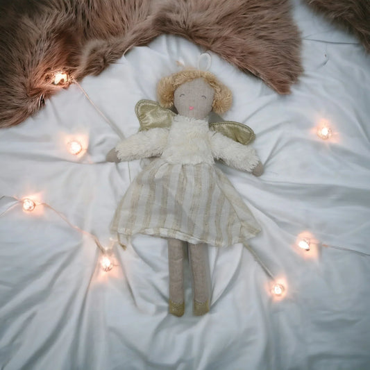 Soft angel doll with gold wings, striped dress, on white bedding with fairy lights and faux fur