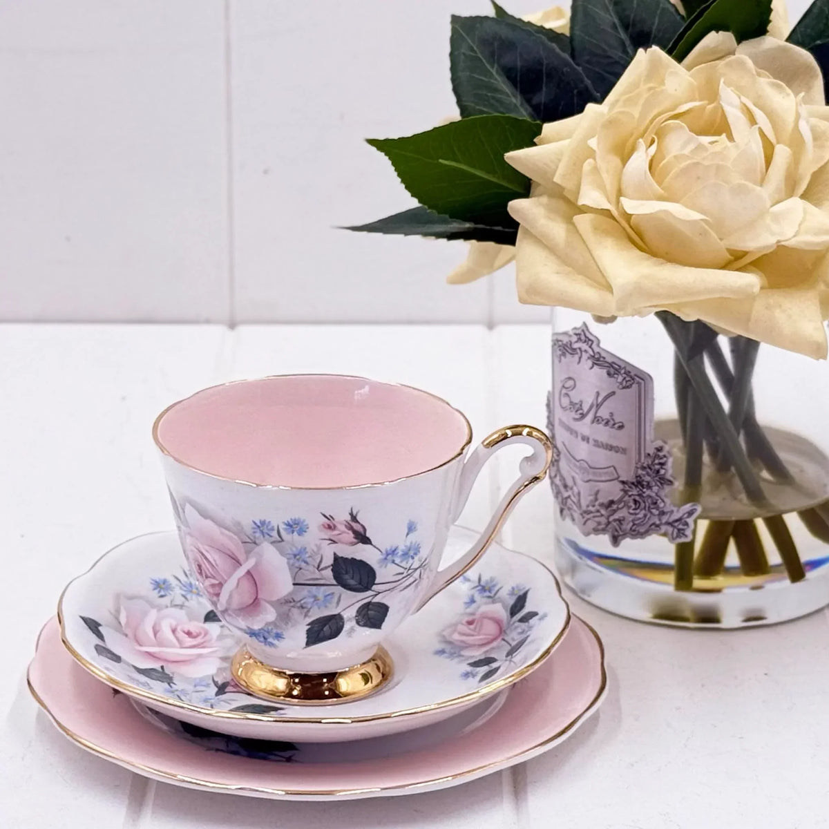 Elegant floral teacup set with pink roses, gold trim, and matching saucer, styled beside a cream rose in vase