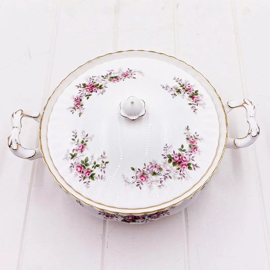 Elegant porcelain tureen with pink rose floral design, gold trim, and decorative handles on white wood background