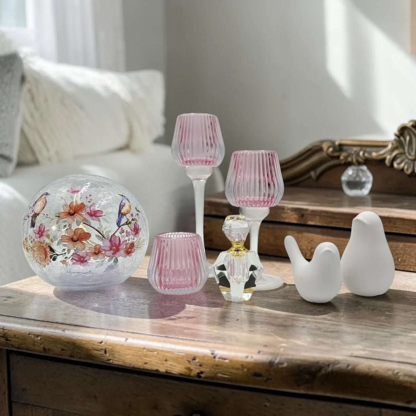 Elegant home decor with floral glass sphere, pink ribbed candle holders, crystal perfume bottle, and white bird figurines on rustic wood table