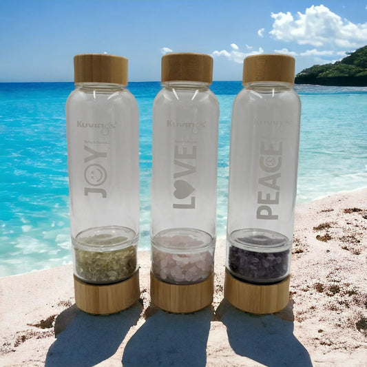 Three glass water bottles with bamboo lids and crystal infusers, labeled Joy, Love, Peace, on a beach.