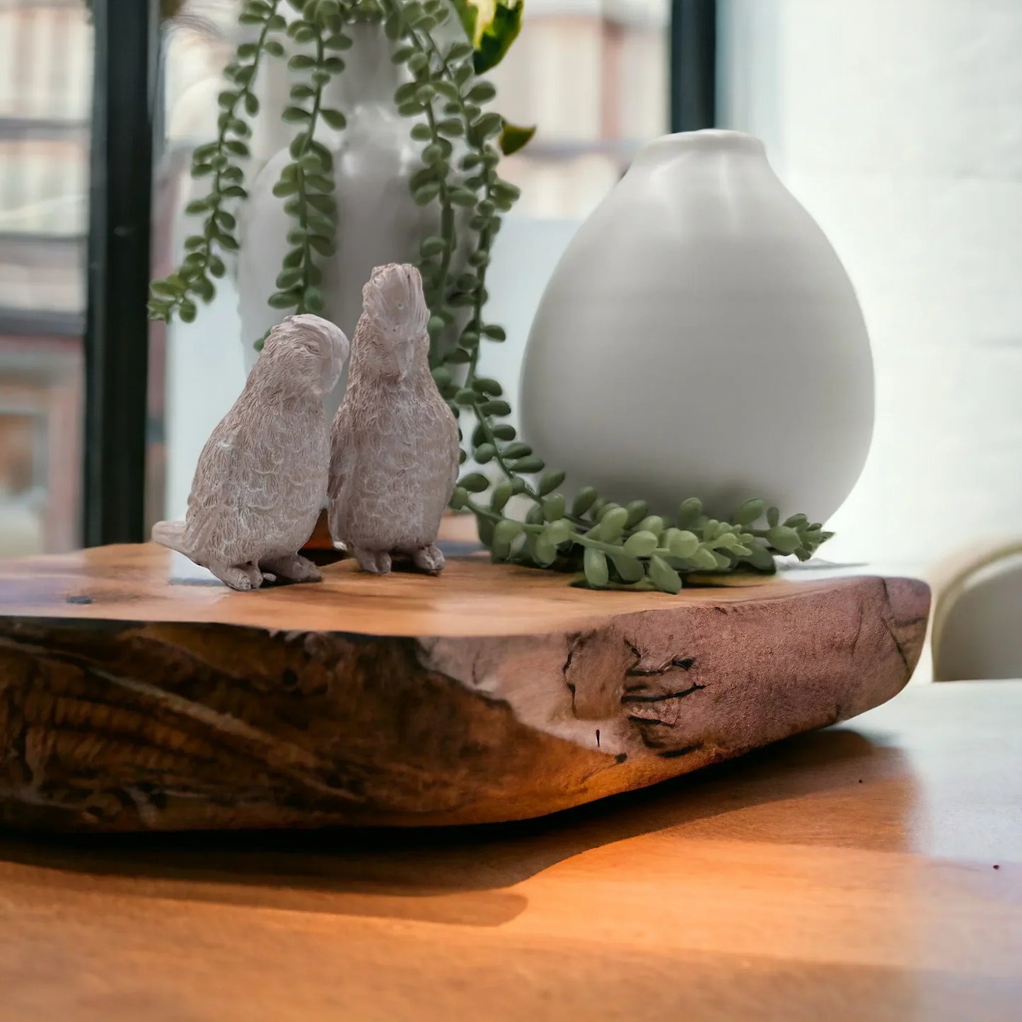 Two textured bird figurines on rustic wood slab with greenery and white vase, home dƩcor accent
