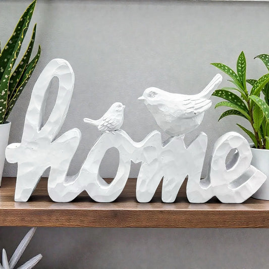 White 'Home' sign with birds, rustic home decor on wooden shelf, flanked by green plants