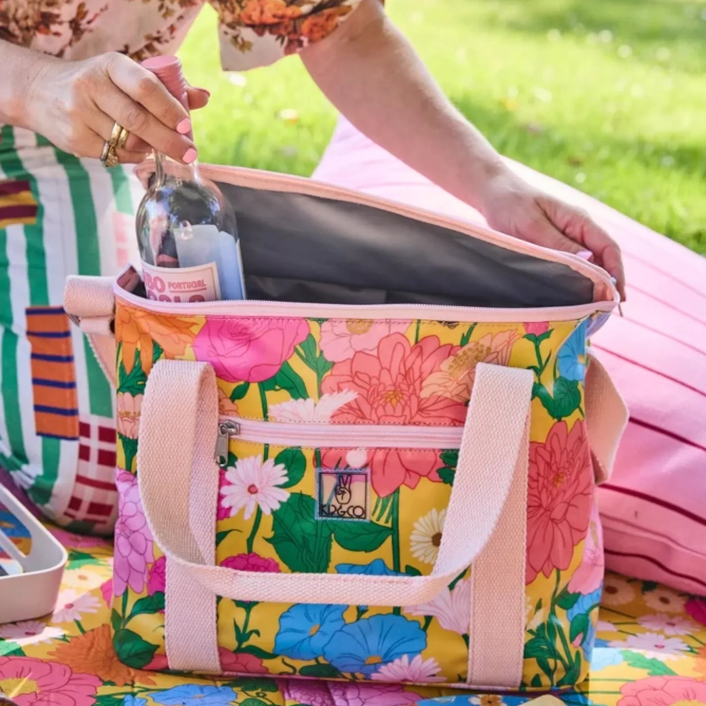 Floral picnic cooler bag with pink handles, holding wine bottle on colorful blanket outdoors