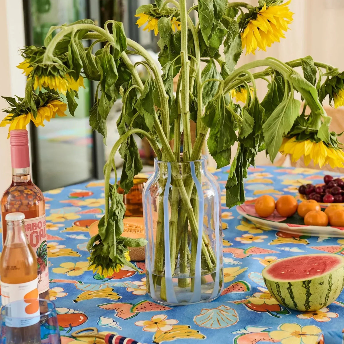 Sunflowers in glass vase on colorful fruit-print tablecloth with watermelon and wine, home décor styling