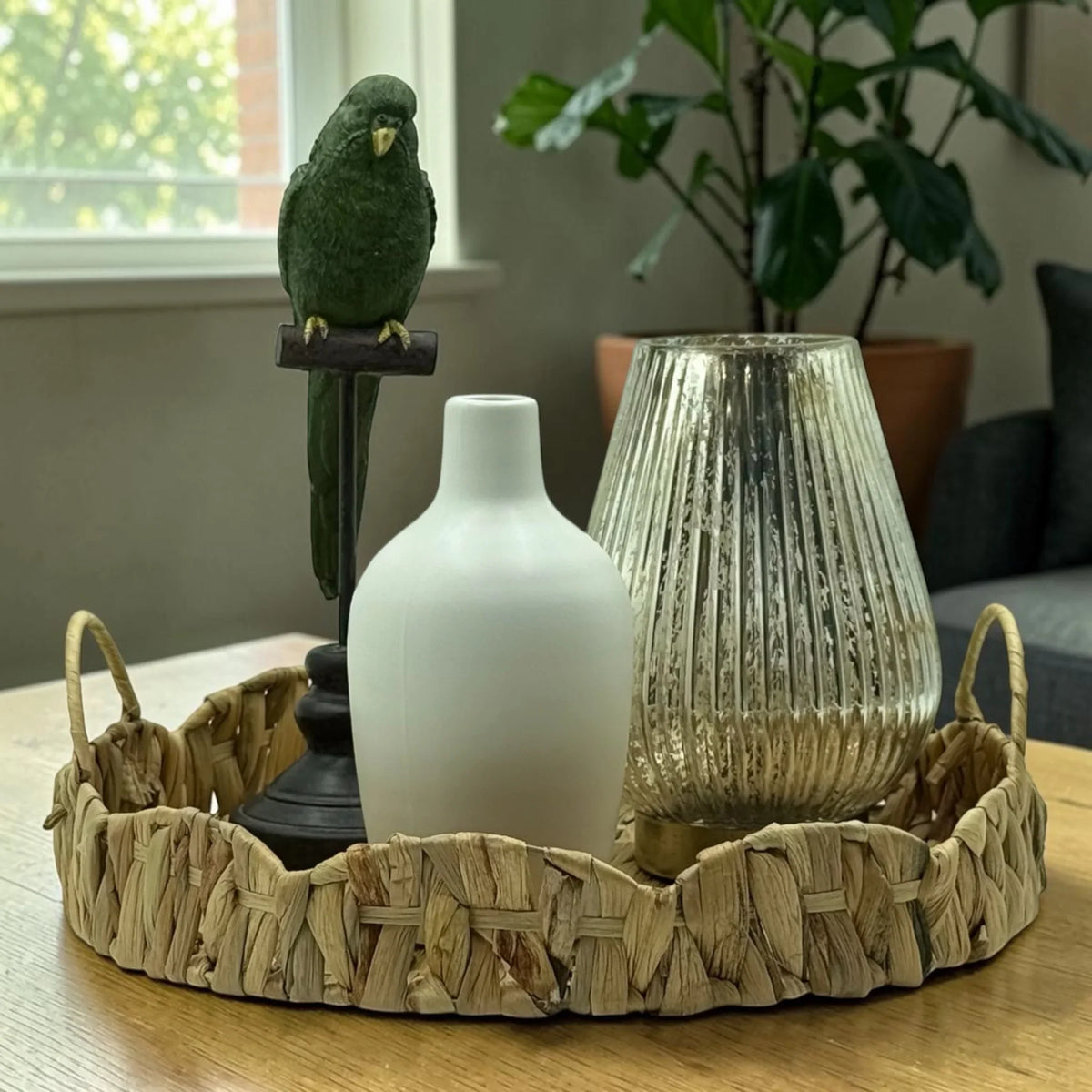 Green parrot figurine, white vase, and mercury glass candle holder on woven tray for home décor
