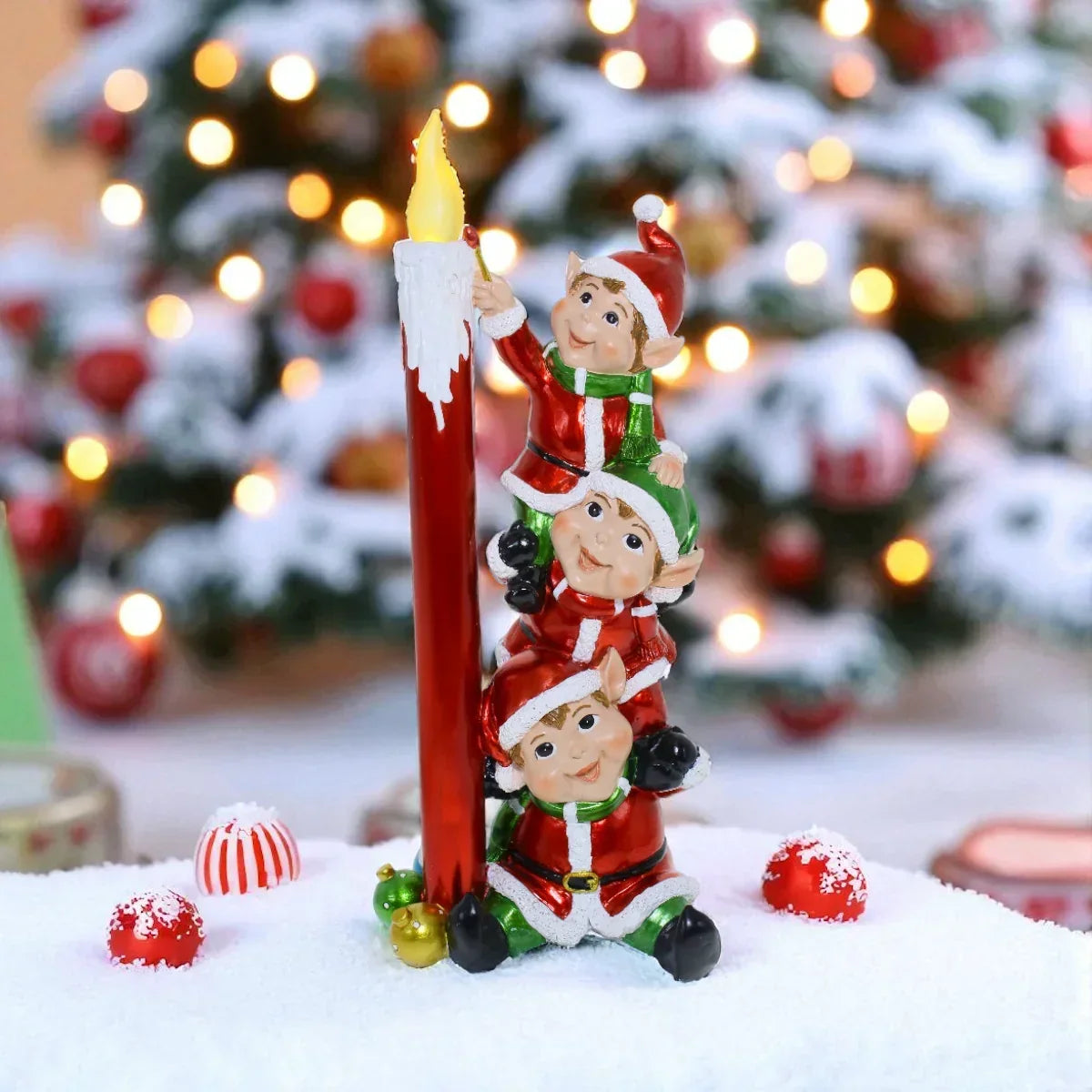 Three festive elves in red and green climbing a tall red candle, surrounded by snowy Christmas ornaments and a lit tree.