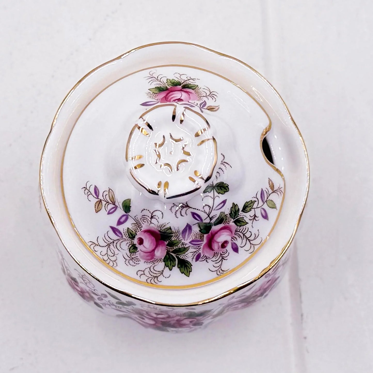 A white Royal Albert  Lavender Rose preserve/sugar/jam pot with a floral design featuring pink roses, green leaves, and gold accents, photographed from a top-down angle on a light textured background.