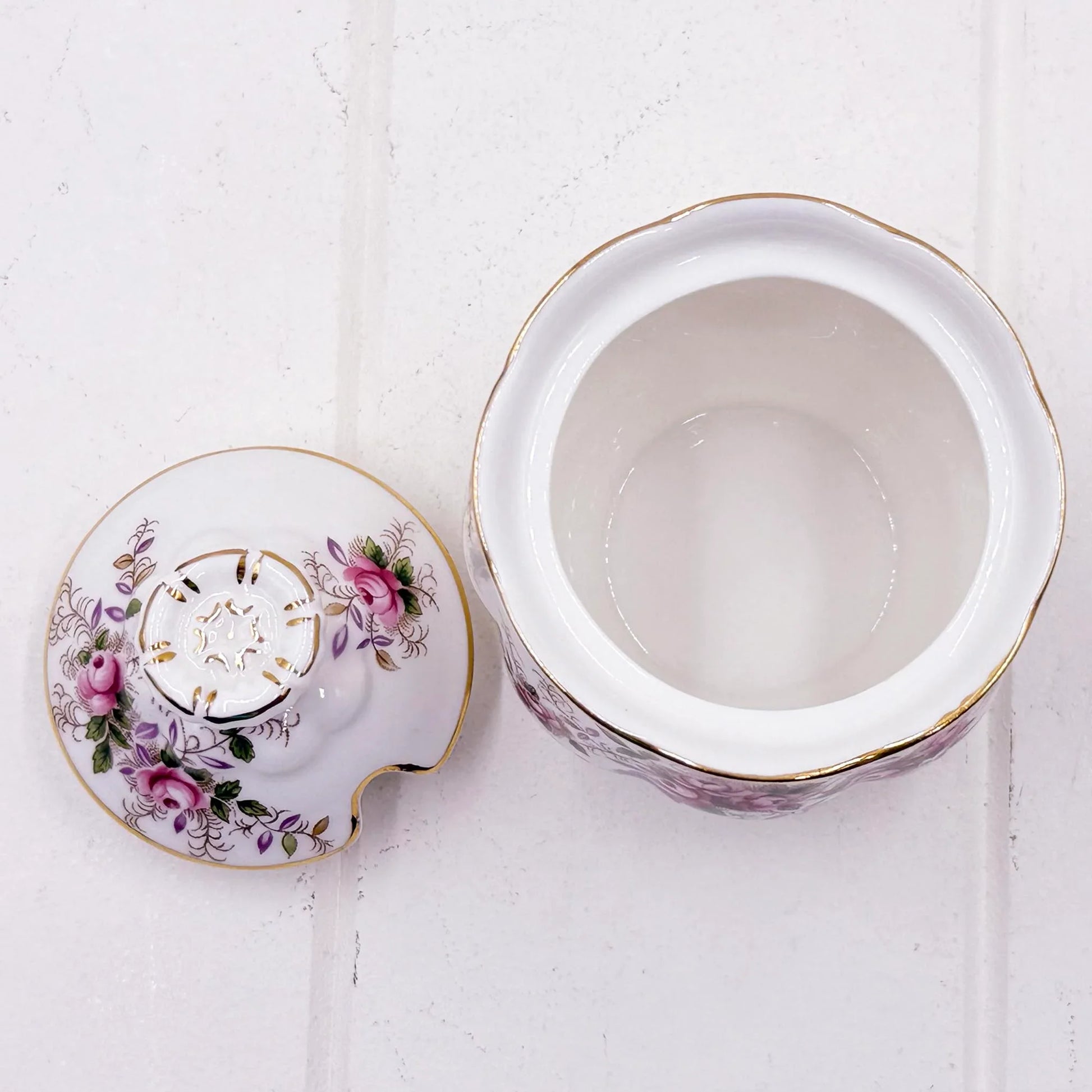 A white Royal Albert lavender Rose bone china sugar pot container with a floral design in pink, purple, and green, accented with gold trim, shown open with its lid placed beside it on a white textured background.