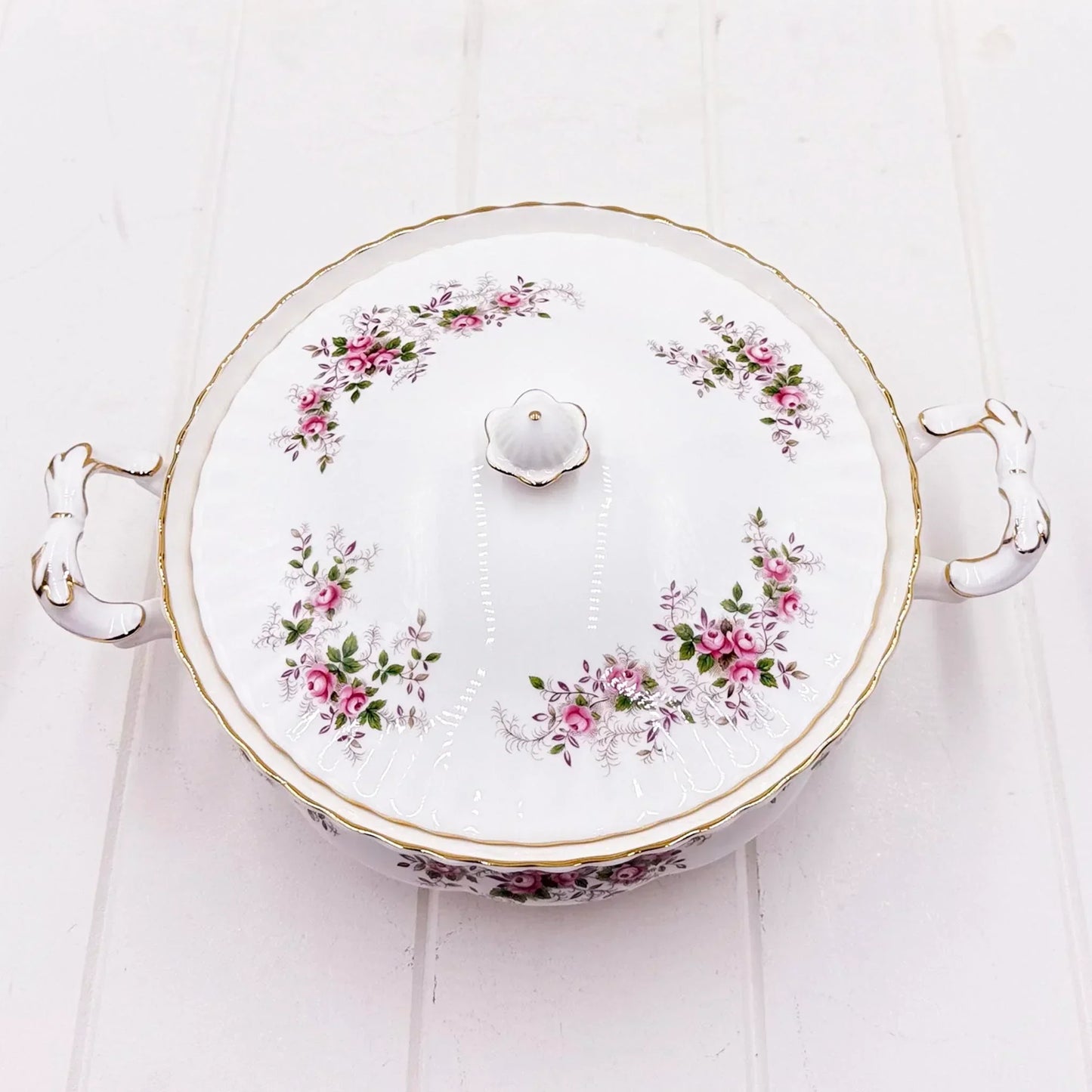 Elegant porcelain tureen with pink rose floral design, gold trim, and decorative handles on white wood background